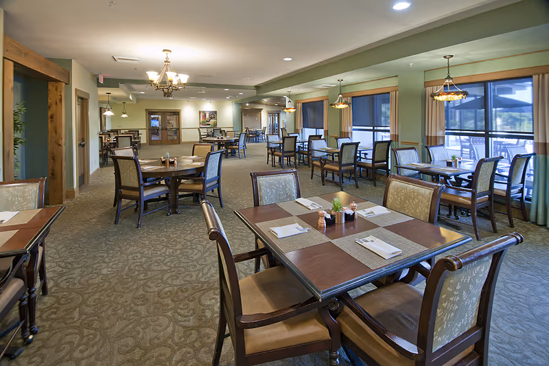 Spacious dining room with multiple wooden tables and chairs set with placemats and napkins and large windows along one side.