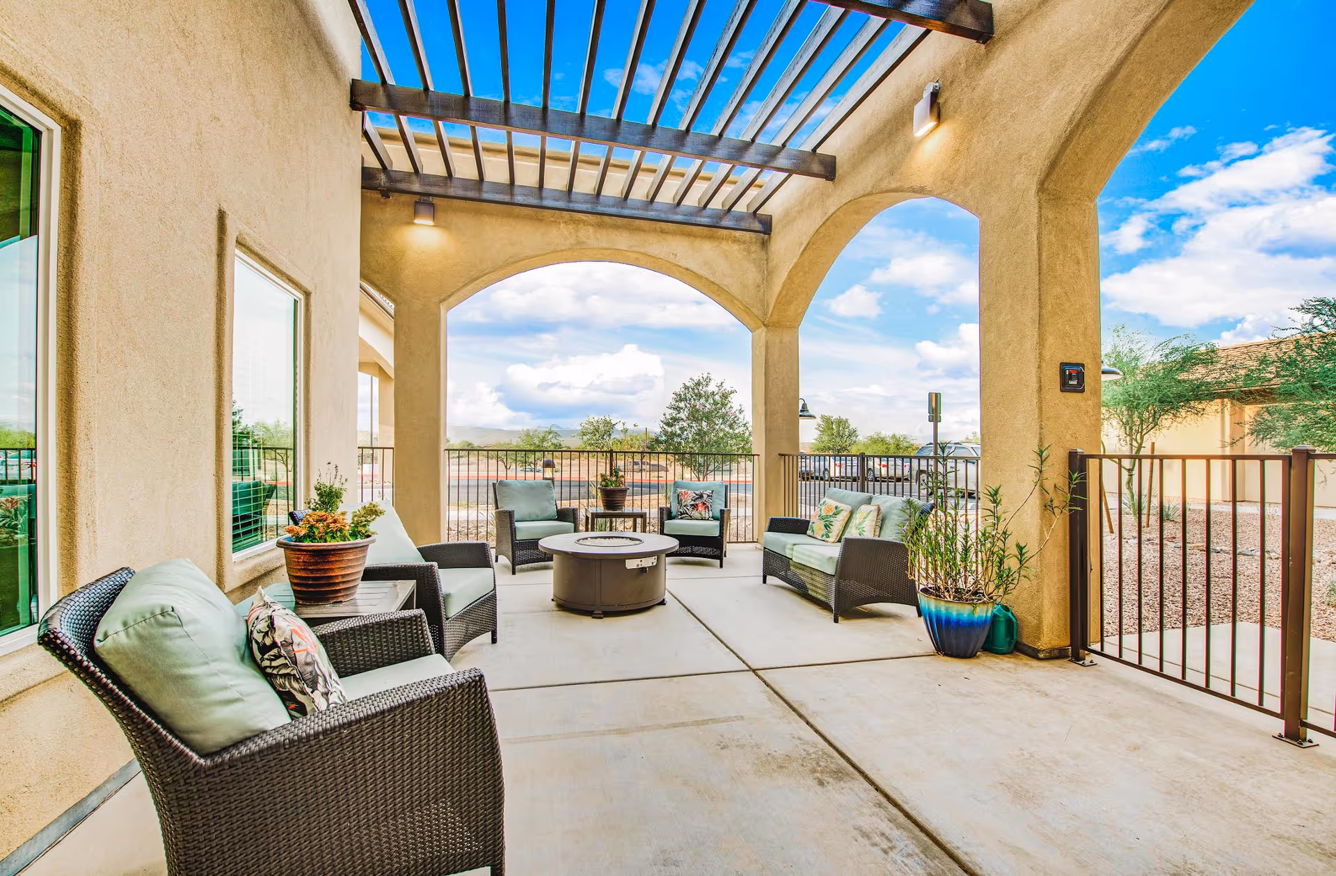 Outdoor covered patio area with wicker chairs and cushioned seating arranged around a circular fire pit table. The patio has beige stucco walls, arched openings, and a pergola overhead. Potted plants are placed around the seating area, and a metal railing encloses the space. The sky is blue with some clouds visible in the background.