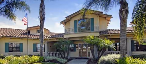 Two-story tan senior living facility exterior with palm trees and an American flag