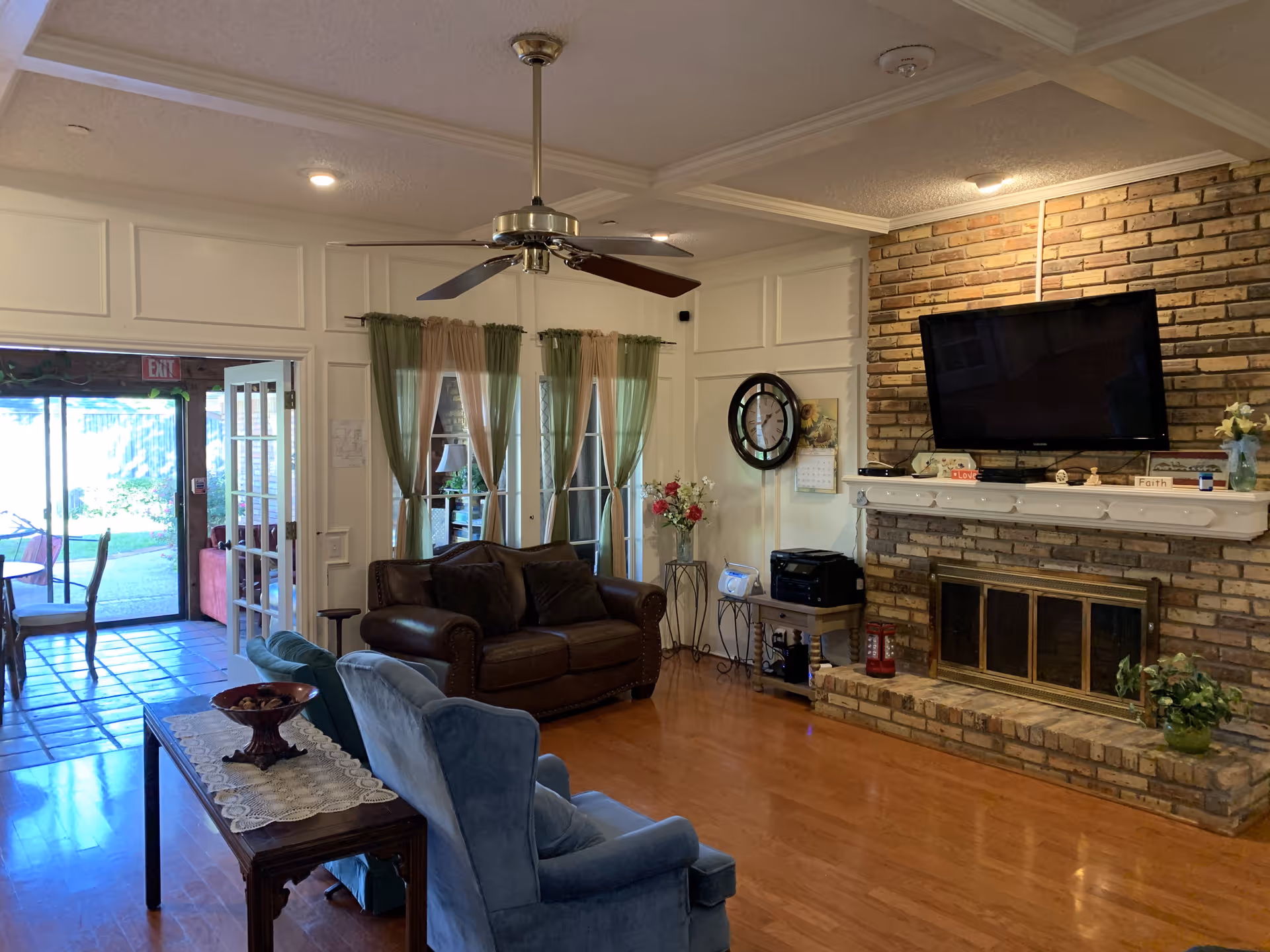 A cozy living room with a brown leather sofa, two blue armchairs, a wooden table with a decorative bowl, a ceiling fan, and a brick fireplace with a mounted flat-screen TV above it. The room has wooden flooring, white walls with paneling, green and beige curtains on glass doors, and a clock on the wall. There is also a small table with a printer and a radio beside the fireplace.