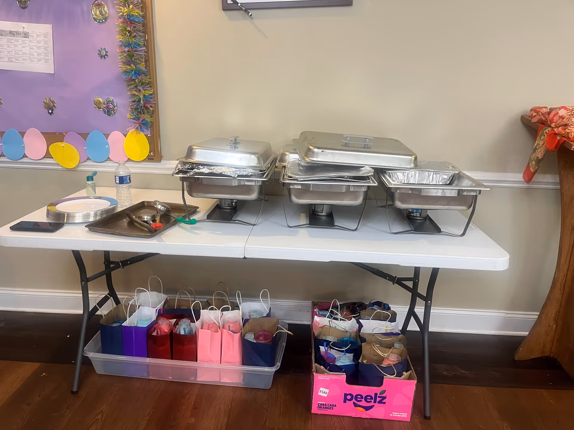 Fold-out table holding three covered chafing dishes, plates and trays with colorful gift bags stored underneath in a decorated room.