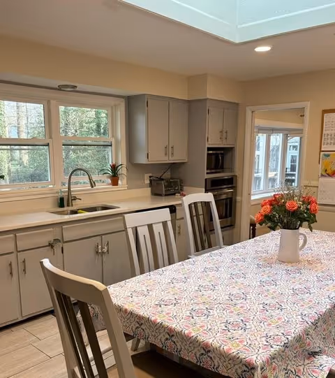 A bright kitchen with white cabinets and a large window above the sink showing greenery outside. A table covered with a patterned tablecloth is in the foreground, with a white vase holding a bouquet of red and orange flowers. There are several white chairs around the table, and a microwave and oven are built into the cabinetry. The room has light-colored tile flooring and a skylight above.