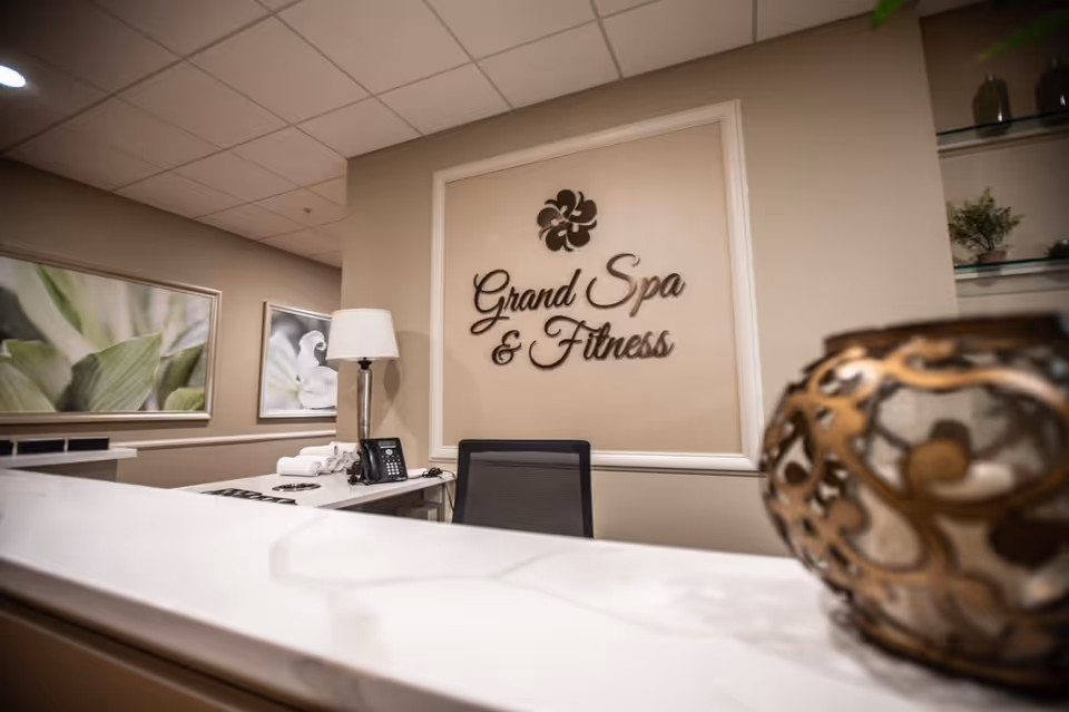 Reception desk inside a spa with a wall sign reading 'Grand Spa & Fitness', a lamp, phone, and decorative vase.