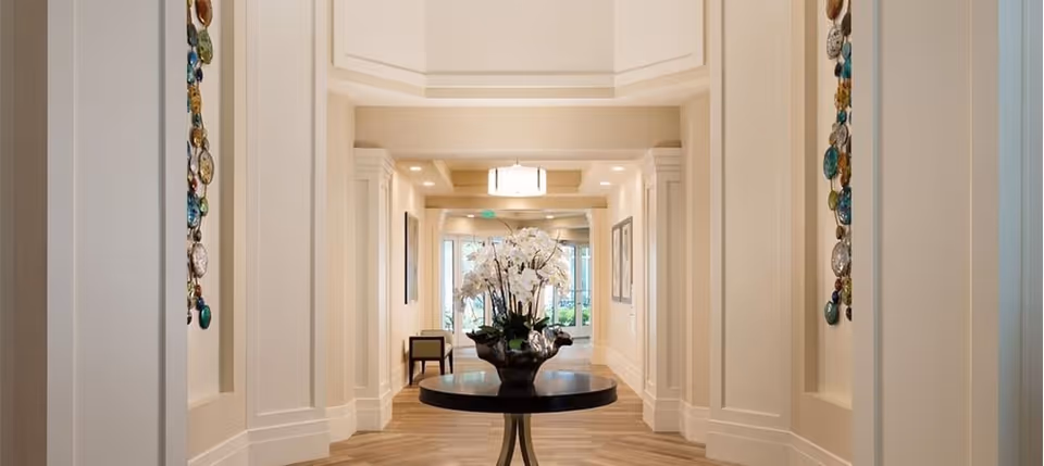 A bright and elegant hallway with cream-colored walls and decorative wall panels. A round table with a large floral arrangement is centered in the hallway. The floor has a light wood pattern, and there are chairs and framed artwork visible further down the hall near glass doors leading outside.