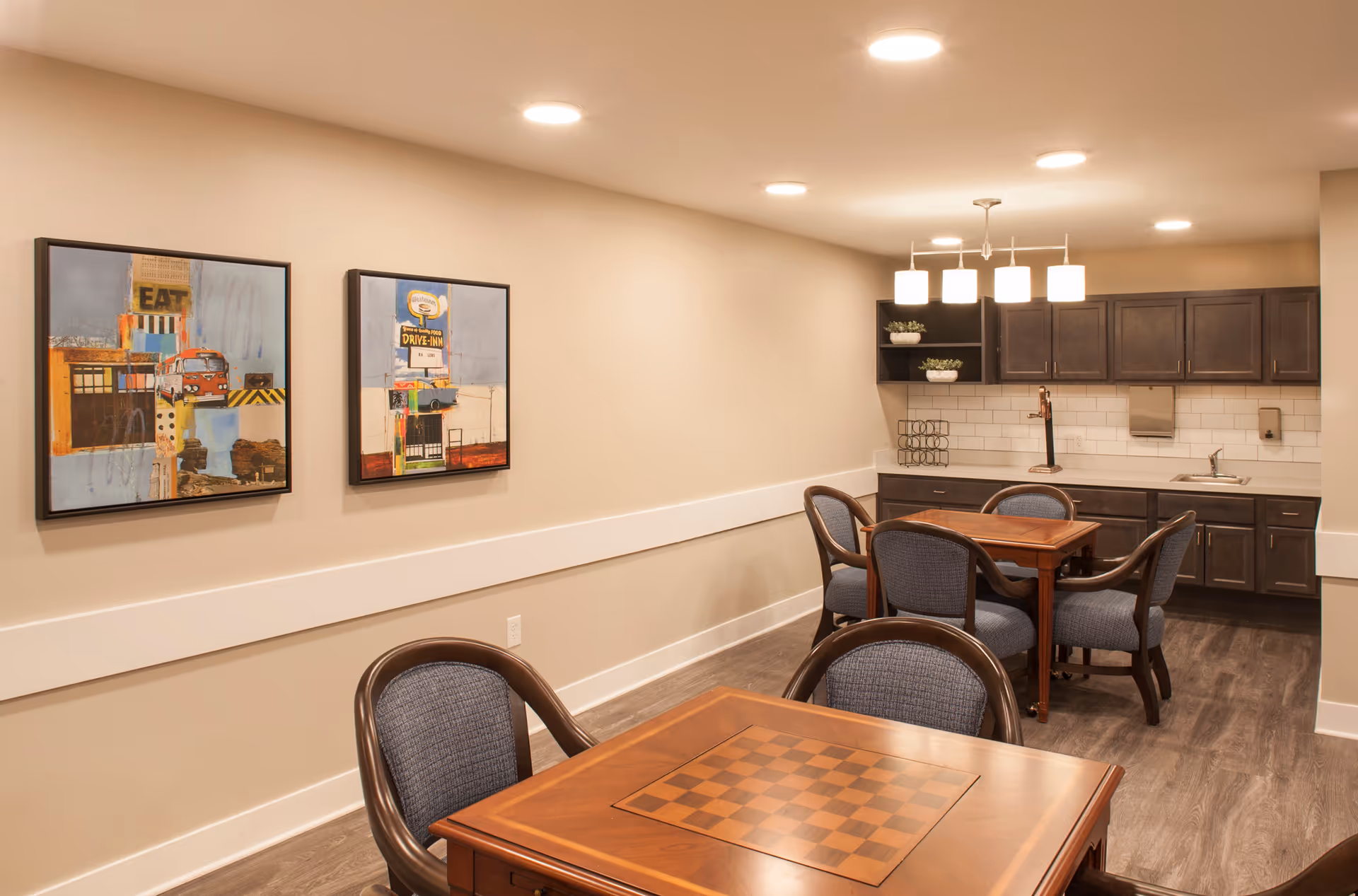 Interior view of a common area with two wooden tables featuring built-in checkerboards, surrounded by cushioned chairs. The room has light beige walls with two colorful framed paintings, dark wood cabinetry with a sink, and modern ceiling lights.