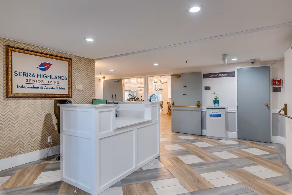Reception area of Serra Highlands Senior Living with a white front desk, a framed sign on the wall displaying the facility name and services, and doors leading to the dining room and storage areas. The floor has a patterned wood and tile design, and the area is well-lit with ceiling lights.
