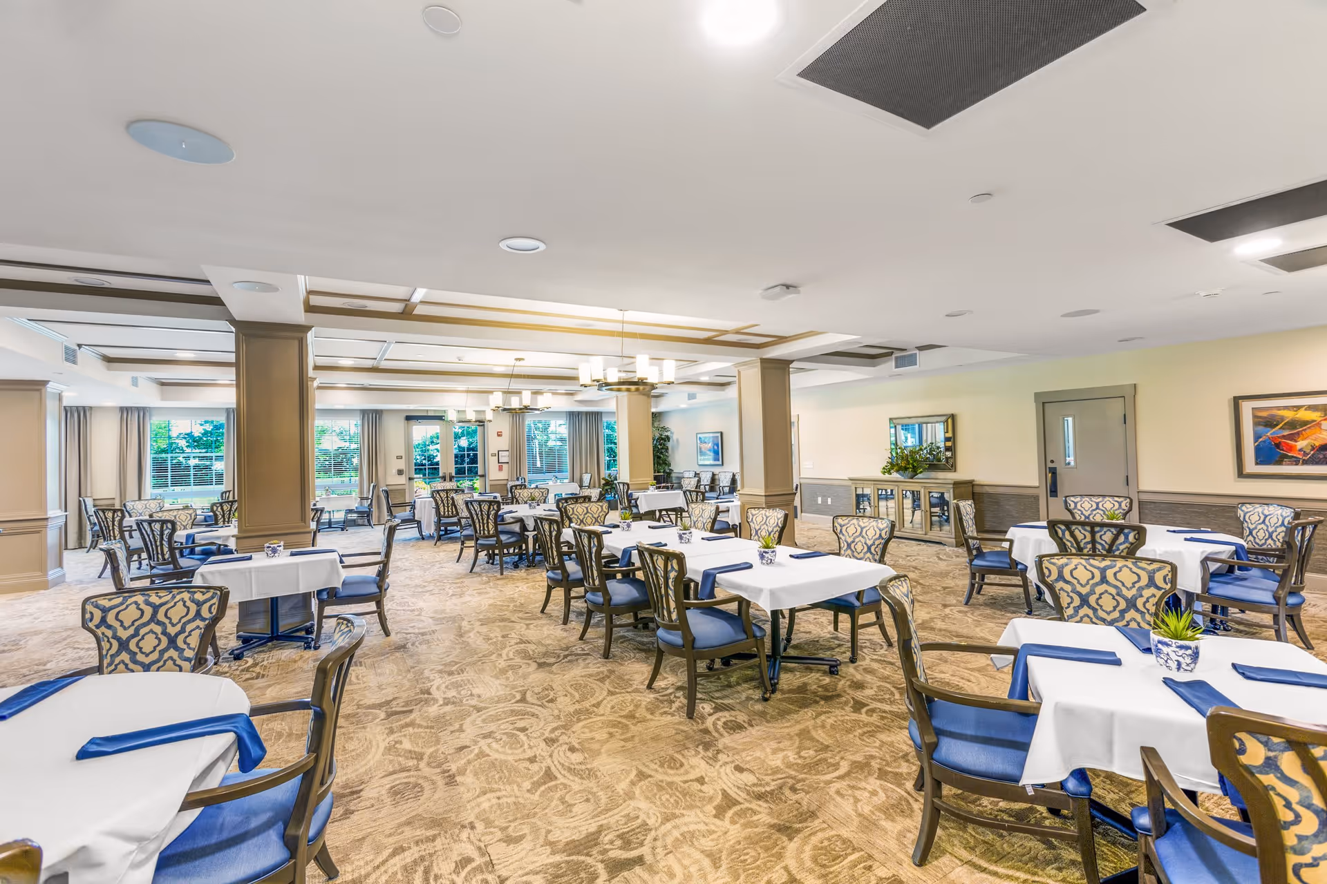 A spacious dining room with multiple tables covered in white tablecloths and blue napkins. Each table is surrounded by patterned chairs with blue cushions. The room has beige walls, carpeted floors with a floral pattern, large windows letting in natural light, and ceiling lights with chandeliers. There are decorative plants and framed artwork on the walls.