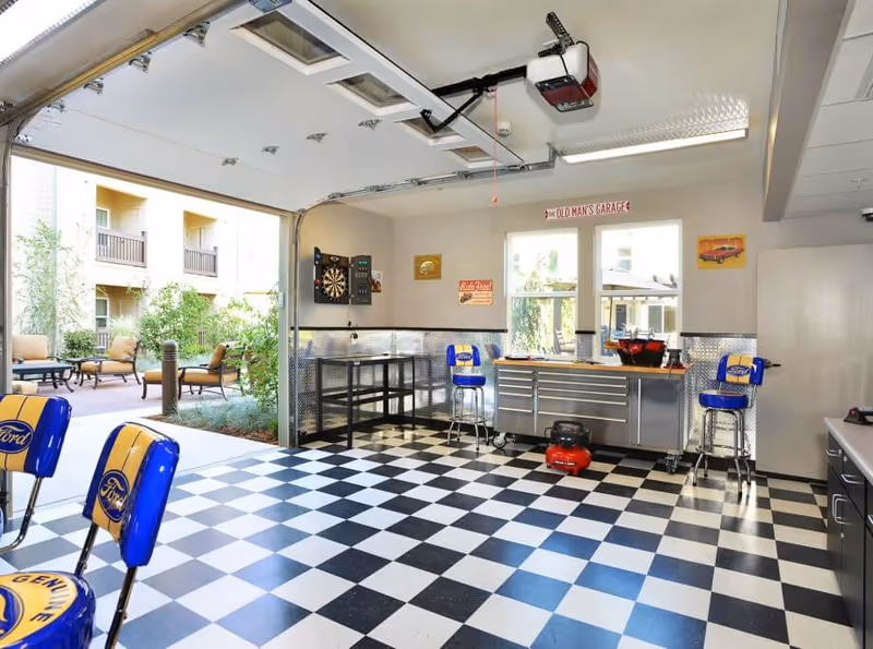 Interior garage-style workshop with a black-and-white checkered floor, metal tool cabinets, stools, and an open door to an outdoor courtyard.