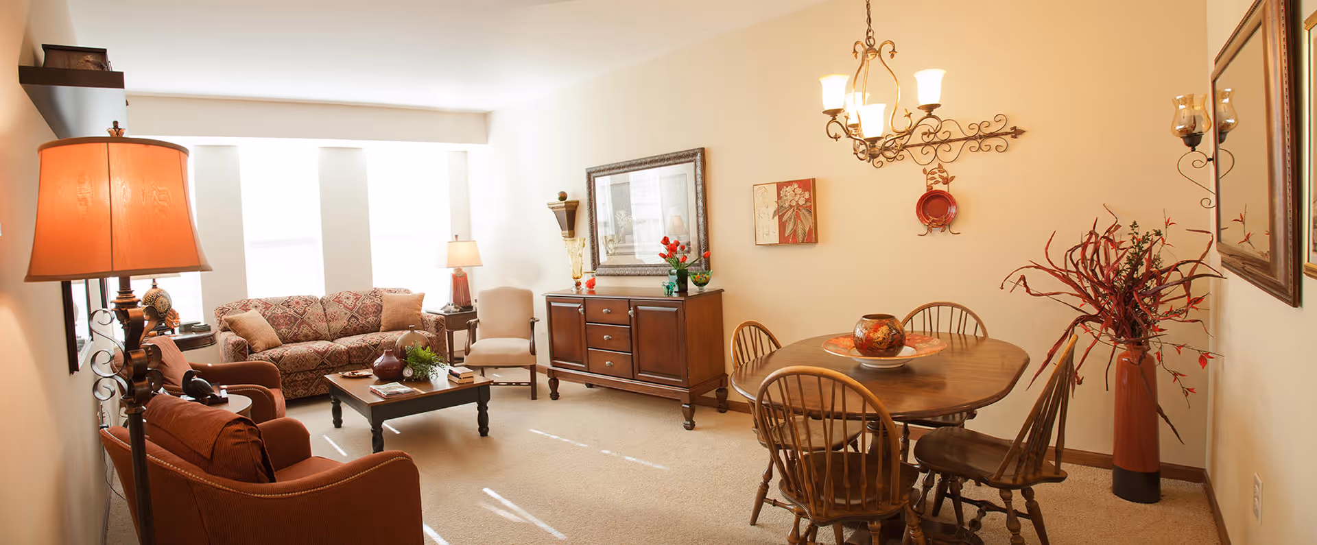 A cozy living and dining area with beige walls and carpet. The living room has a patterned sofa, two armchairs, a coffee table with decorative items, and two lamps providing warm lighting. The dining area features a wooden round table with four chairs, a chandelier above, and decorative wall art and a tall vase with branches.