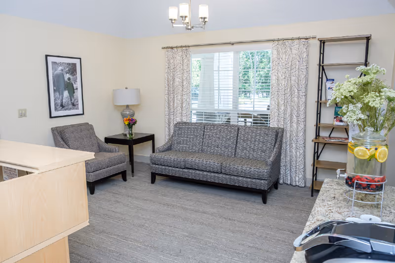 A cozy living room area with a patterned gray sofa and matching armchair, a side table with a lamp and flowers, a framed black and white photo on the wall, large window with patterned curtains, a wooden shelf with brochures, and a countertop with a glass dispenser of water with lemon and berries.