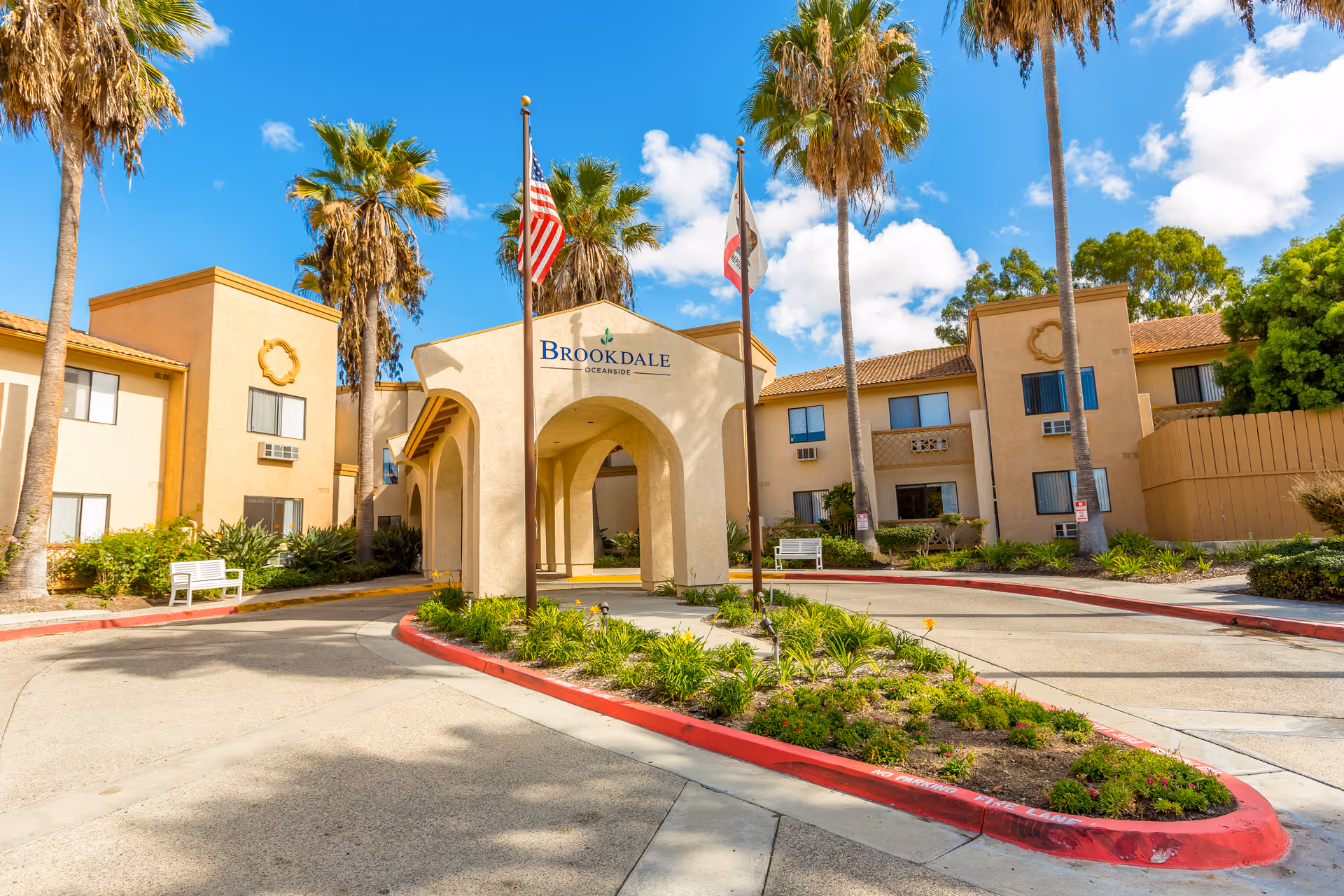 Exterior view of Brookdale Senior Living facility in Oceanside with a beige stucco building, palm trees, two flagpoles with American and California state flags, and a circular driveway with landscaped greenery in the center.