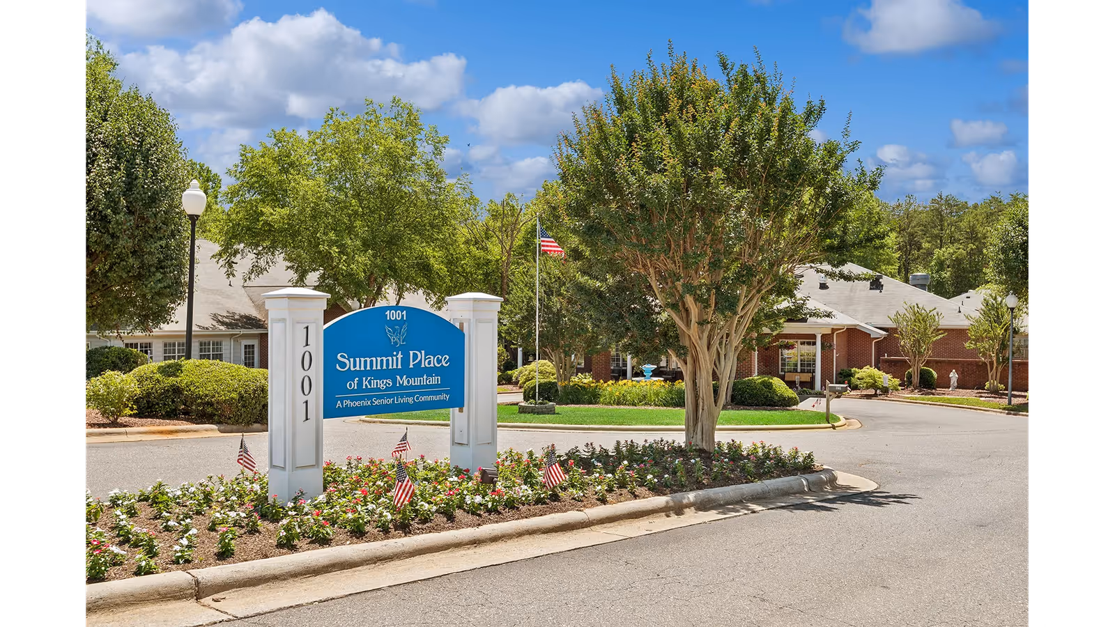 Entrance area of Summit Place of Kings Mountain senior living community with a blue sign displaying the facility name, surrounded by landscaped flower beds with small American flags, trees, and a clear blue sky with some clouds.