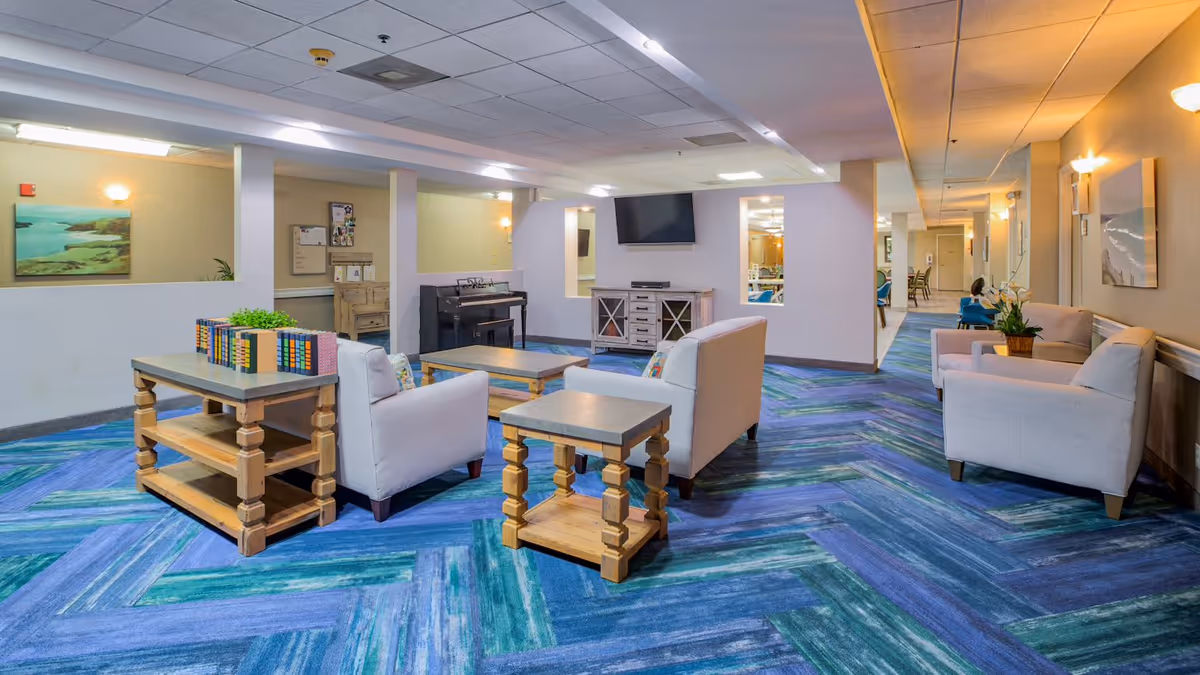 A spacious and brightly lit common area in an assisted living facility featuring blue and green patterned carpet, light-colored armchairs and sofas arranged around wooden tables, a piano, a wall-mounted flat screen TV, and artwork on the walls.