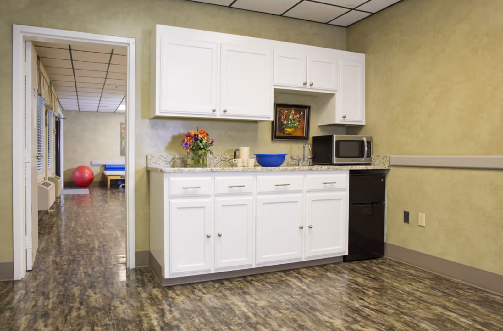 A small kitchenette area with white cabinets, a granite countertop, a microwave, a small black refrigerator, a blue bowl, a vase with colorful flowers, and a framed floral painting on the wall. The floor has a marbled pattern, and there is an open doorway leading to another room with a red exercise ball and a blue padded table.