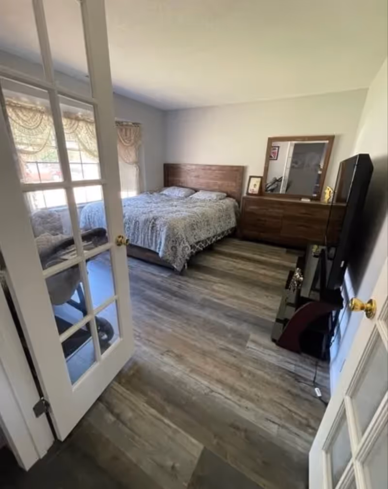 Bright bedroom with a wooden bed and dresser, a TV on a stand, and wood-look flooring seen through glass-paneled French doors.