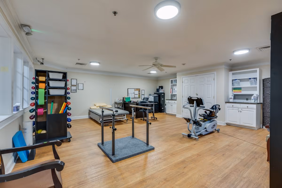 A spacious therapy or exercise room with wooden flooring, featuring a stationary exercise bike, parallel bars for walking support, a treatment table with pillows, a desk with office supplies, and shelves with various exercise equipment and supplies. The room has white walls, ceiling lights, a ceiling fan, and large windows letting in natural light.
