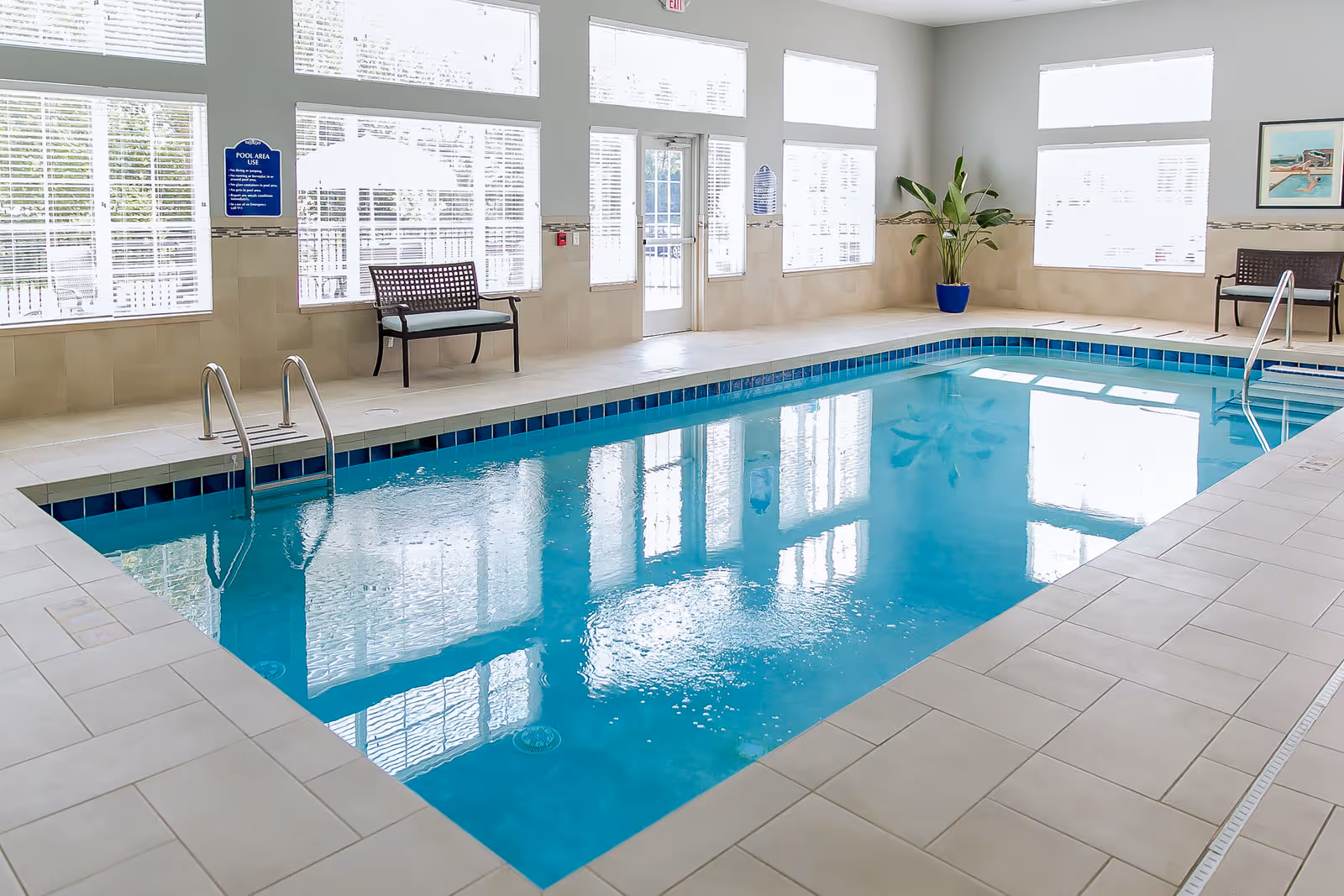 Indoor swimming pool with tiled deck, metal handrails, benches, large windows, and a potted plant.