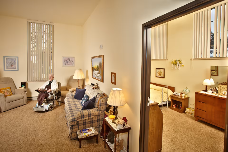 A cozy living room area in a senior living facility with an elderly woman sitting on a mobility scooter. The room features a plaid sofa with cushions, an armchair, a side table with a lamp, and framed artwork on the walls. Adjacent to the living room is a bedroom with a bed, nightstand, dresser, and lamps, visible through an open doorway.
