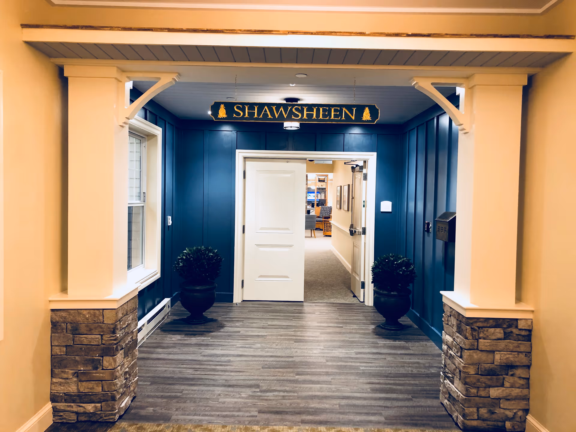 Interior hallway entrance with blue paneled walls and a sign above the door that reads 'SHAWSHEEN'. The hallway has wood-look flooring, two potted plants on either side of the door, and a white double door leading to another room with chairs and framed pictures visible inside.