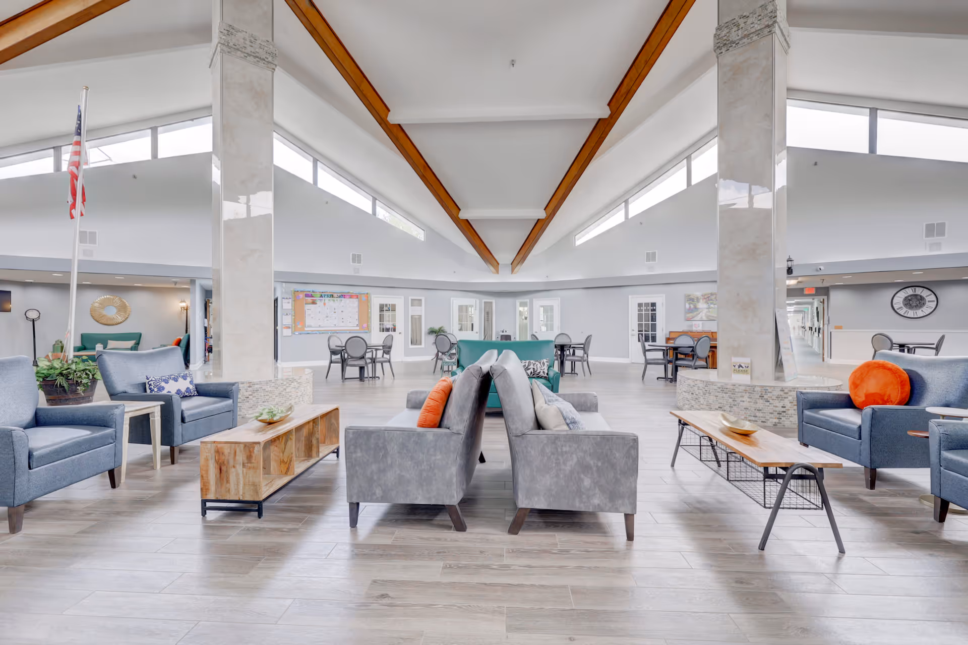 A spacious and bright senior living facility common area with modern furniture including gray and blue armchairs, gray sofas with orange and patterned pillows, wooden coffee tables, and several small round tables with chairs. The room features high ceilings with wooden beams, large windows near the ceiling allowing natural light, two large marble pillars, and an American flag on a pole. The walls are light gray with white trim, and there is a bulletin board and a large clock visible in the background.