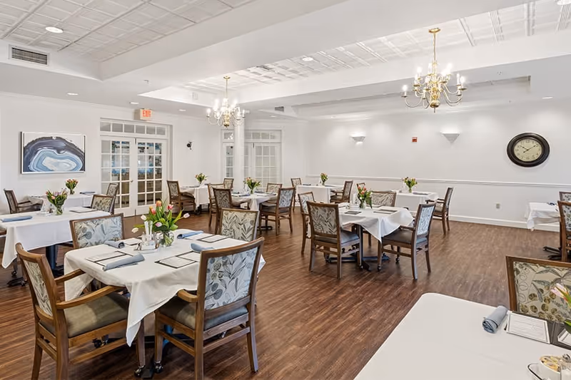 A bright and spacious dining room with multiple tables covered in white tablecloths, each set with menus, napkins, and small flower arrangements. The room features wooden chairs with patterned upholstery, hardwood floors, white walls, a clock on the wall, and two chandeliers hanging from a coffered ceiling. French doors and a piece of abstract wall art are visible in the background.