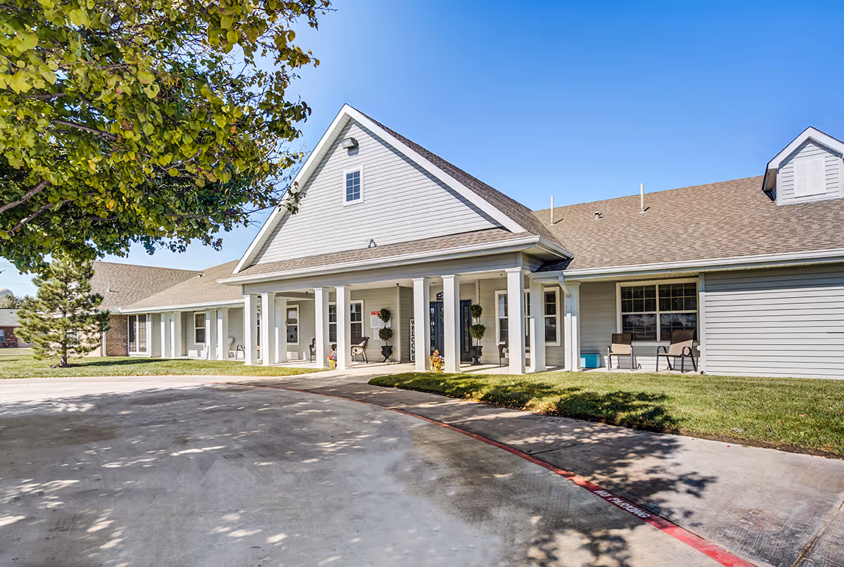 Exterior view of Meredith Court, a single-story building with a gabled roof and white siding. The entrance features a covered porch supported by white columns, with chairs and potted plants near the doors. There is a tree on the left side and a clear blue sky above.