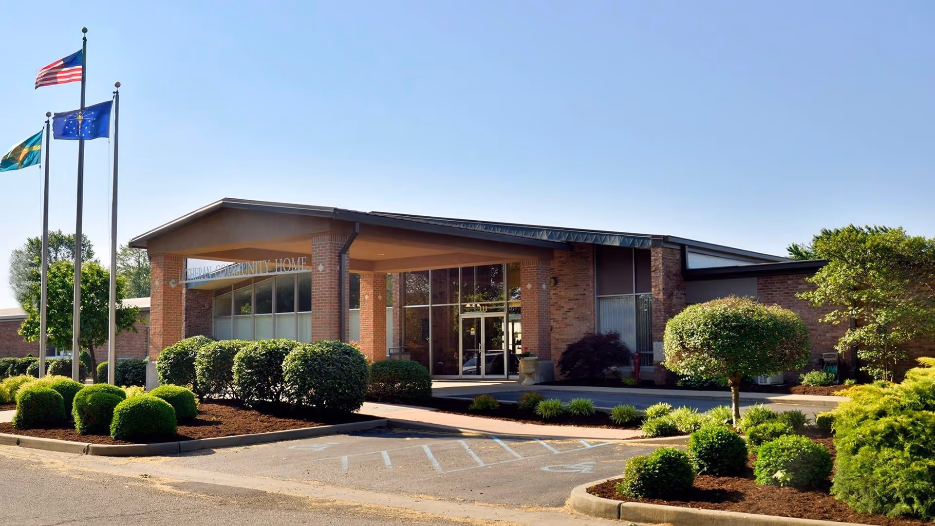 Exterior view of Lutheran Community Home, Inc. building with a covered entrance, surrounded by neatly trimmed bushes and trees. Three flagpoles with flags are visible on the left side under a clear blue sky.