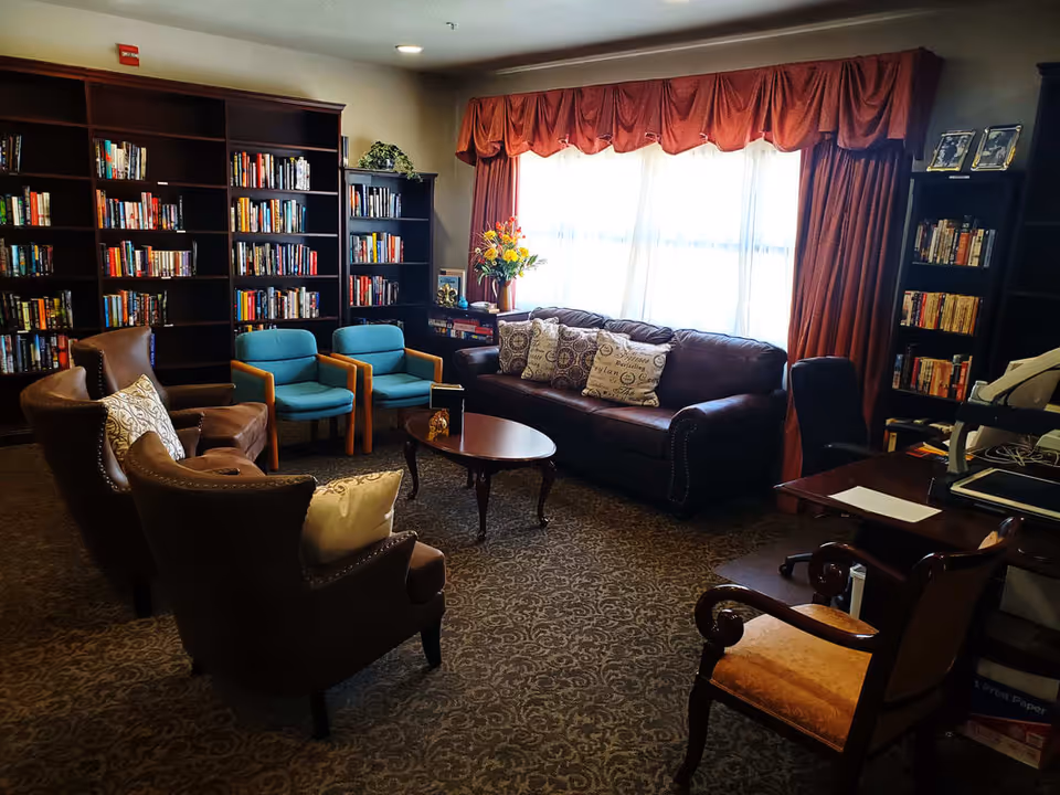 A cozy library or lounge area with bookshelves filled with books along the walls, a brown leather sofa with decorative pillows, two blue armchairs, and two brown leather armchairs arranged around a wooden coffee table. There is a desk with a chair and office equipment on the right side, and a large window with red curtains letting in natural light.