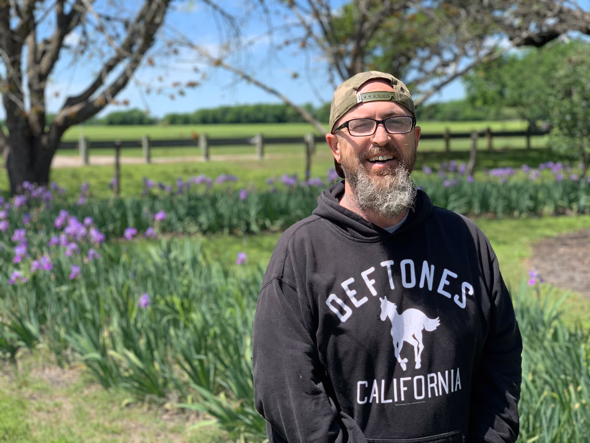 A man wearing glasses, a backwards cap, and a black hoodie with the words 'DEFTONES CALIFORNIA' stands outdoors in front of a garden with purple flowers and a wooden fence, with trees and a clear blue sky in the background.