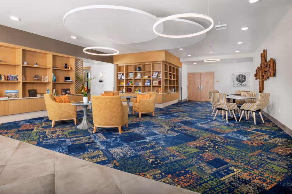 A spacious senior living common area with colorful patterned carpet, four mustard-yellow armchairs around a small round table with a plant, and a set of striped chairs around a round table near the wall. The room features wooden bookshelves filled with books and decorative items, modern circular ceiling lights, and a wall decoration with a cinema poster.