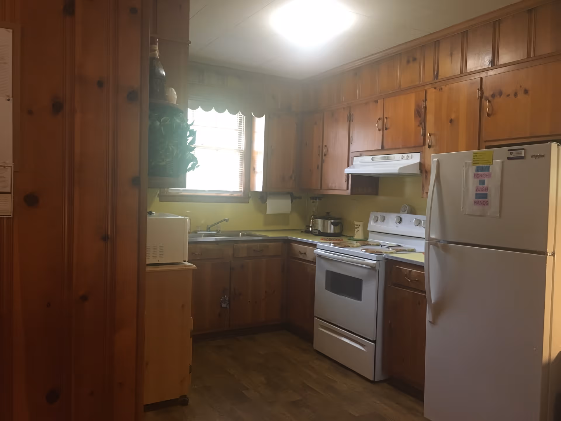A kitchen with wooden cabinets, a white stove with an oven, a white refrigerator with a sign on it, a microwave on a small wooden stand, a window with blinds and a scalloped valance, and a countertop with a slow cooker and other kitchen items.