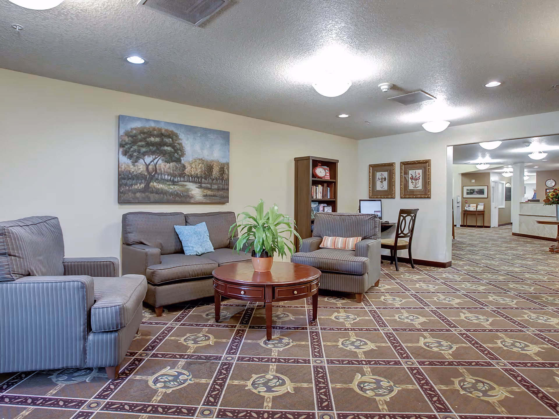 A cozy seating area in a senior living facility with two armchairs and a loveseat arranged around a round wooden coffee table with a potted plant. The walls are decorated with a large painting of a tree and two framed floral prints. There is a bookshelf with books and a clock, and a desk with a chair. The floor has a patterned carpet, and the ceiling has recessed lighting and ceiling fixtures. The space opens into a hallway with more furniture and decorations visible.