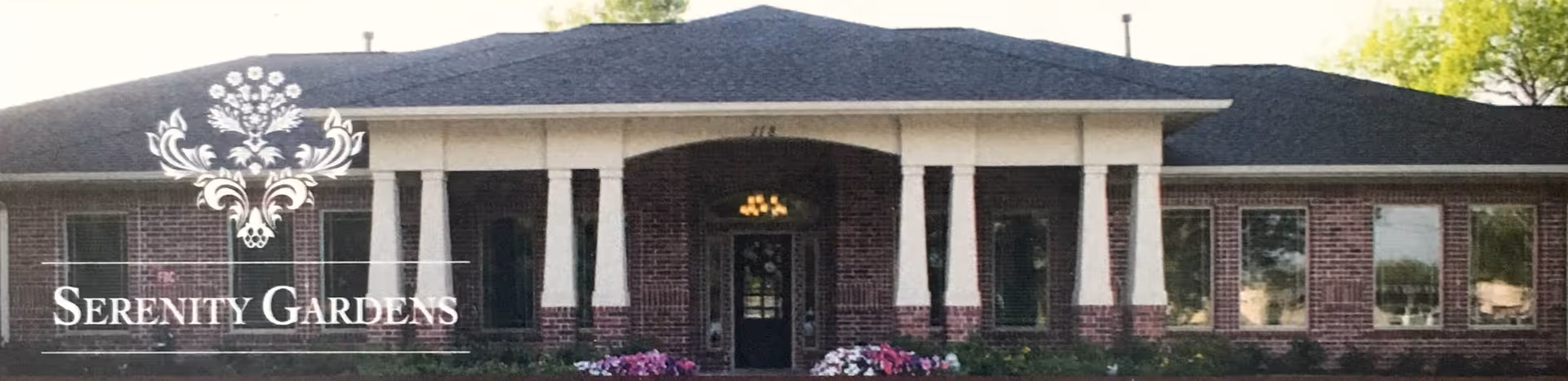 Single-story brick building with a columned front entrance, flower beds, and an overlaid "Serenity Gardens" logo.