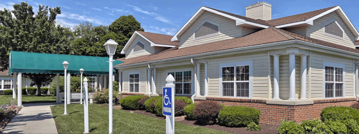 Exterior view of a single-story building with beige siding and a brown roof, surrounded by green bushes and trees. A sidewalk leads to a covered entrance with a green awning supported by white columns. Several white lamp posts line the walkway, and a blue handicap parking sign is visible near the front.