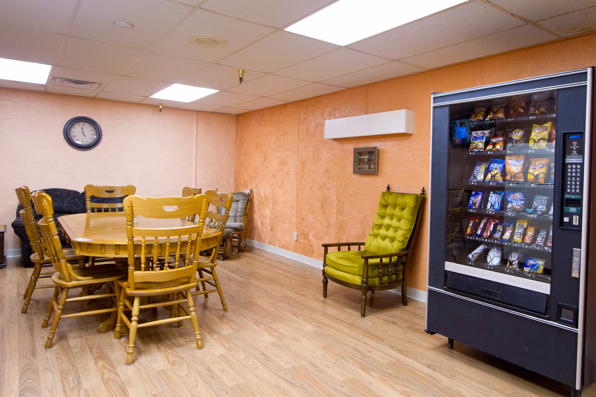 A room with a wooden dining table surrounded by wooden chairs, a green cushioned armchair against an orange textured wall, a vending machine filled with snacks, a clock on a beige wall, and a wooden floor.