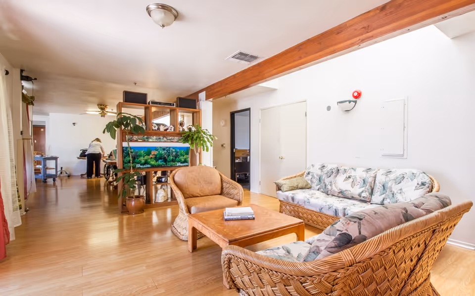A bright and spacious living room area with wicker furniture including a sofa and two chairs with floral cushions, a wooden coffee table with books on it, and a wooden shelving unit with a large aquarium and potted plants. The room has light wood flooring, white walls, and a wooden ceiling beam. In the background, a person is seen attending to something near a table.