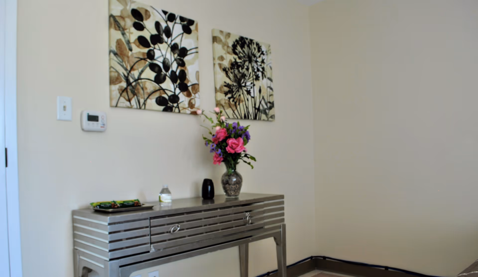 A corner of a room with a metallic console table against a beige wall. On the table, there is a vase with pink and purple flowers, a small black object, a water bottle, and a tray with some items. Above the table, two framed artworks featuring botanical designs hang on the wall. A thermostat is mounted on the wall to the left.