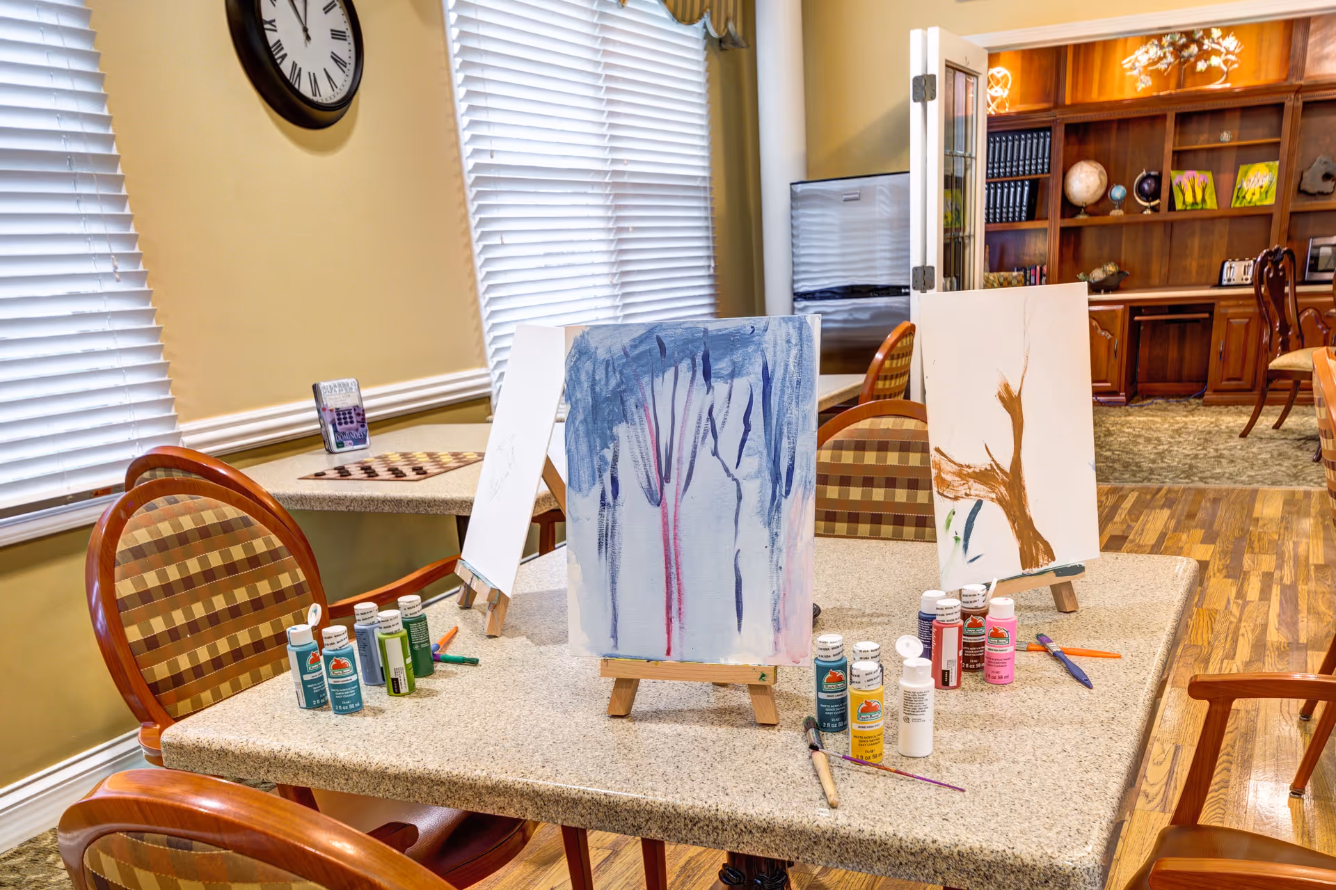 A well-lit activity room with a table set up for painting, featuring three canvases on small easels with partially completed paintings. Various bottles of paint and paintbrushes are scattered on the table. The room has wooden chairs with patterned cushions, large windows with blinds, a clock on the wall, and a bookshelf with decorative items in the background.