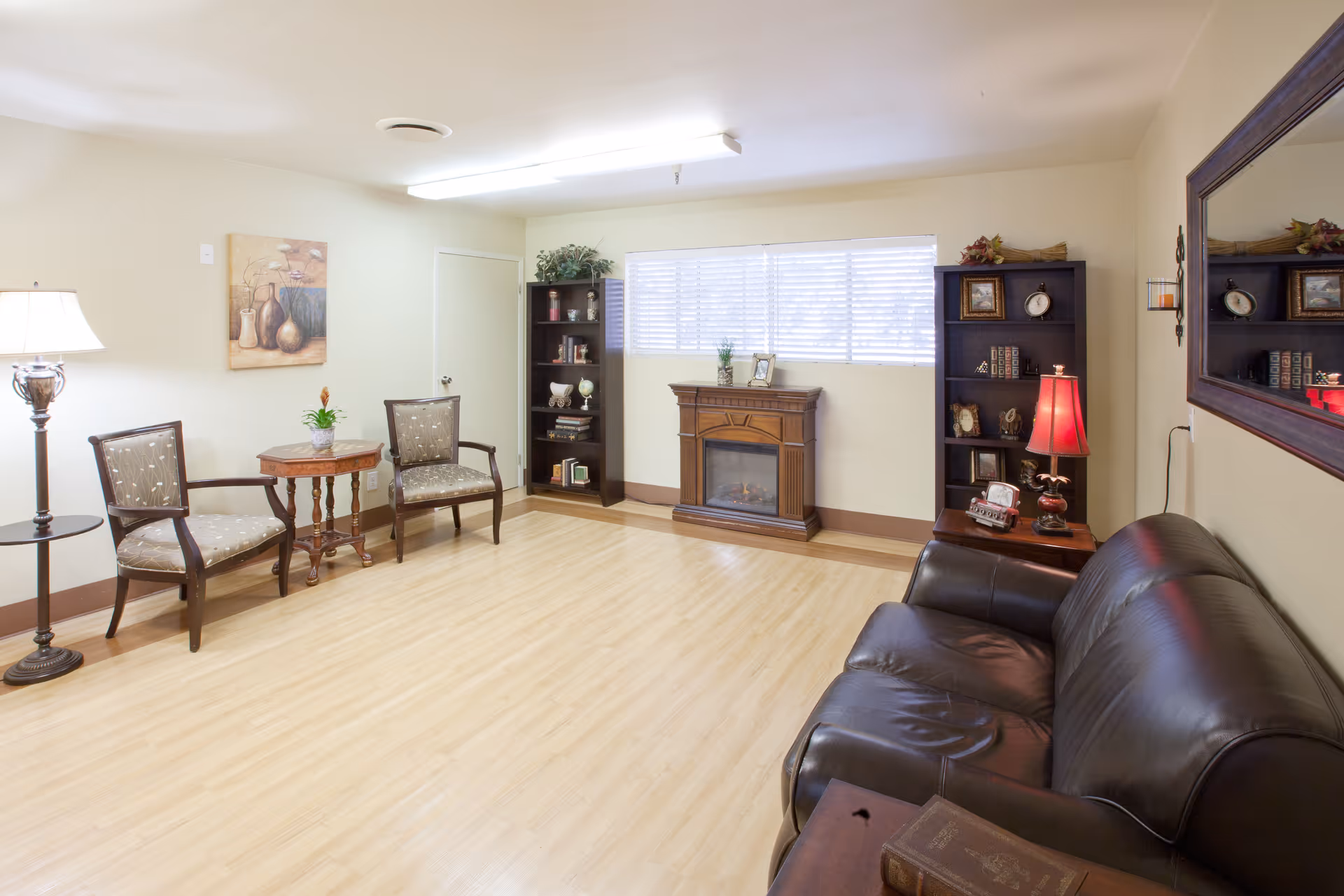 A cozy living room with light wood flooring, a black leather sofa, two wooden chairs with patterned cushions, a small round wooden table with a plant, two dark wood bookshelves filled with decorative items, a wooden electric fireplace, a large mirror on the wall, and a floor lamp and table lamp providing warm lighting.
