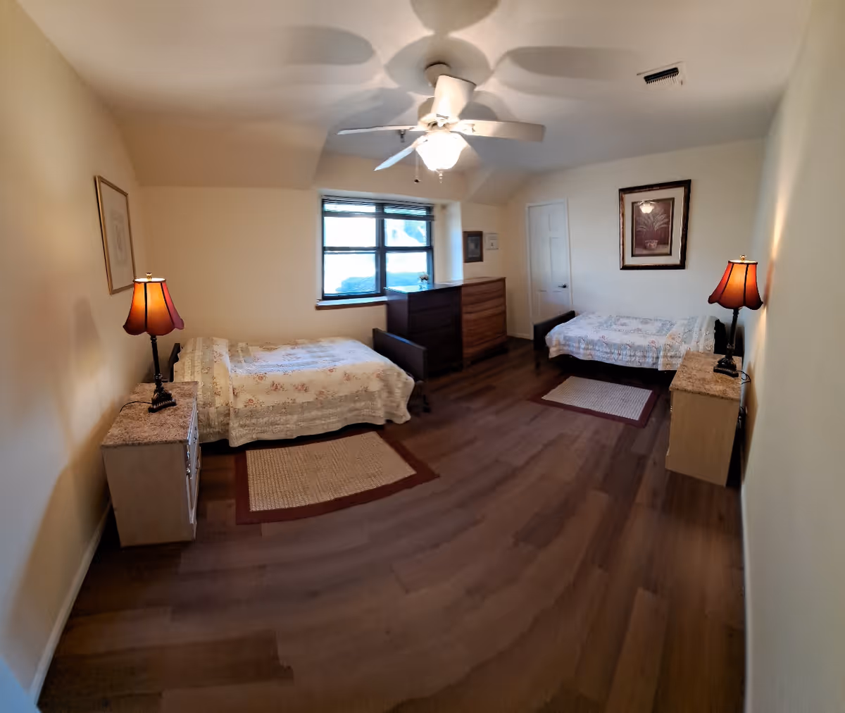 A double-bedroom in a senior living facility with two beds, dressers, lamps, and ceiling fan.