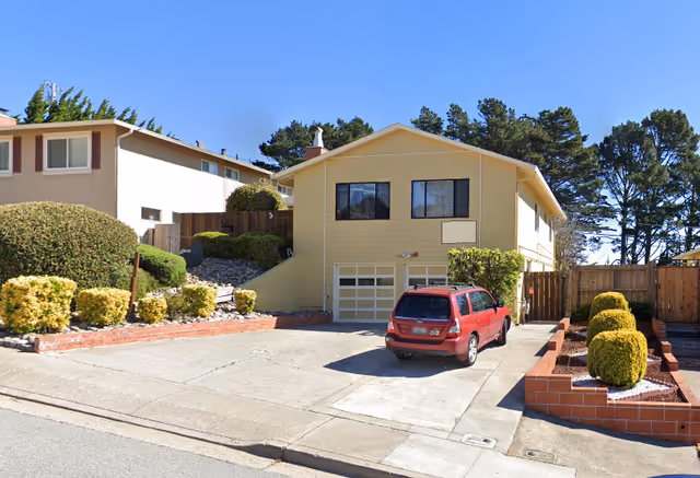 Exterior view of a two-story yellow house with a driveway and a red station wagon parked in front. The house has two large windows on the upper floor and a garage door below. There are neatly trimmed bushes and a brick retaining wall along the driveway. Trees and neighboring houses are visible in the background under a clear blue sky.