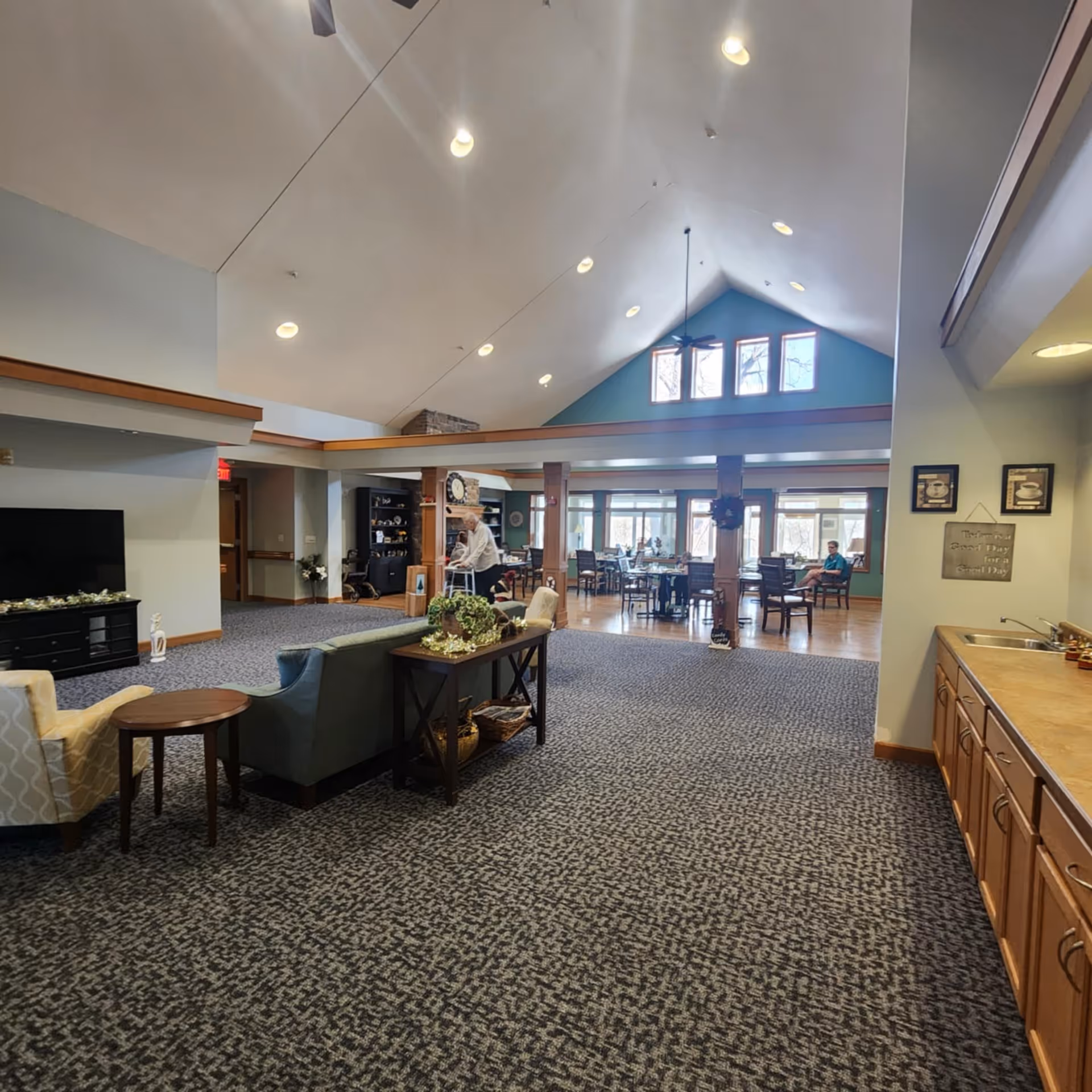 Spacious common area in an assisted living facility with high vaulted ceilings and recessed lighting. The room features a seating area with chairs and a couch, a television on a stand, and a kitchenette with a sink and cabinets on the right. In the background, there is a dining area with tables and chairs near large windows letting in natural light. Two elderly individuals are visible, one using a walker and another seated at a table.