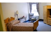 A simple elderly care facility bedroom with a single wooden bed covered with a patterned bedspread, a bedside table with a lamp and flowers, a comfortable armchair near a window with floral curtains, and a wooden dresser with multiple drawers.