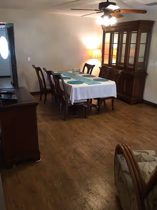 A dining room with a wooden table covered by a white tablecloth and six chairs around it. The table is set with blue placemats and a glass pitcher. There is a wooden china cabinet with glass doors against the wall, a lit table lamp beside it, and a ceiling fan with lights above the table. The floor is wooden, and there is a doorway leading to another room with a door that has an oval window.