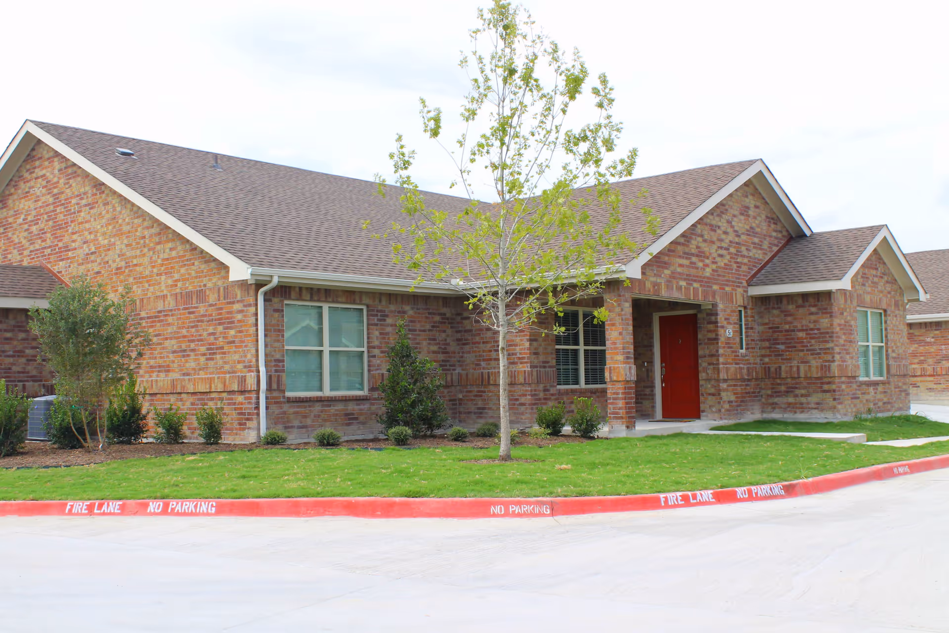 Exterior view of a single-story brick building with a red door, several windows, a small tree, and landscaped bushes in front. The building has a sloped roof and is surrounded by a concrete driveway with red-painted curbs marked with 'FIRE LANE NO PARKING'.
