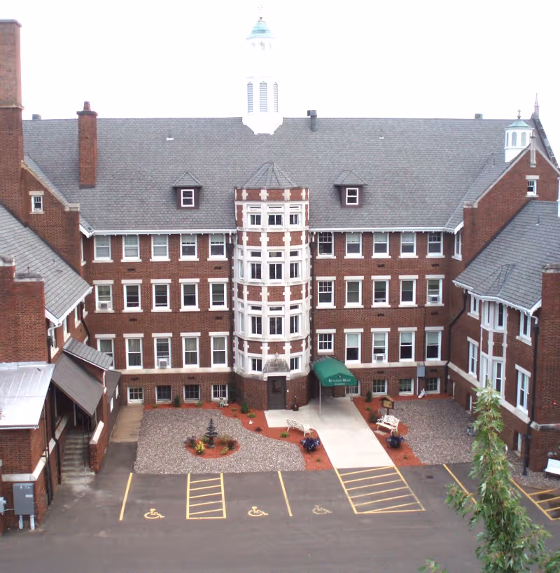 Exterior view of a large brick building with multiple windows and a central tower-like structure. The building has a gray shingled roof and a small courtyard area with benches, a fountain, and landscaped plants. There are several handicap parking spaces in front of the building entrance, which is covered by a green awning labeled 'Rutledge Home'.