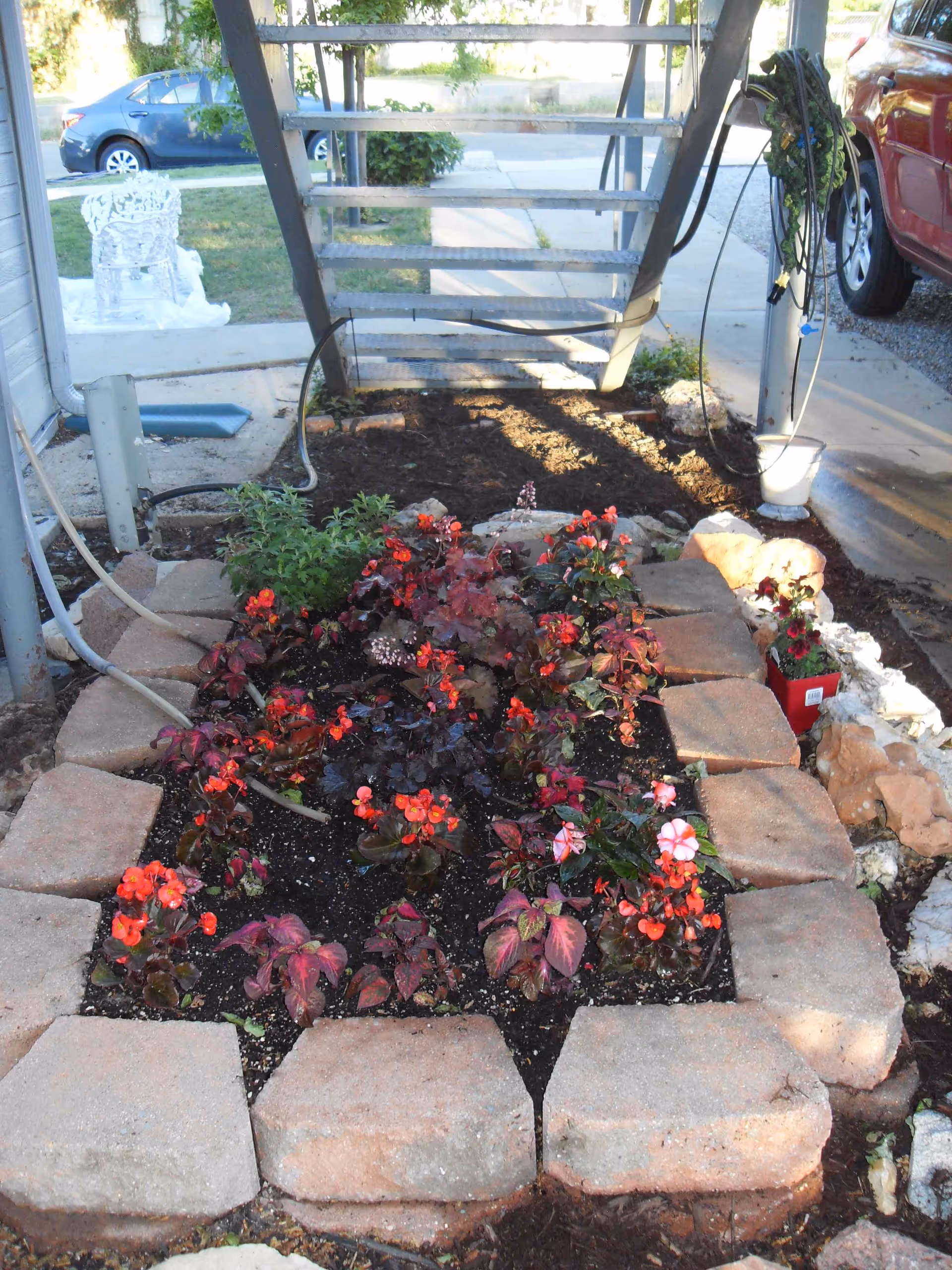 A small garden bed bordered by large rectangular stones, filled with various flowering plants with red and pink blooms. The garden is located under an outdoor metal staircase with a concrete walkway and parked cars visible in the background.