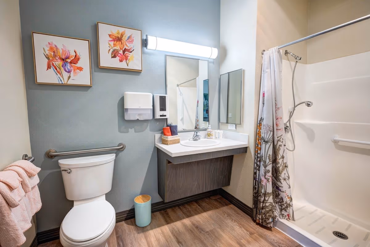 A clean and modern bathroom featuring a white toilet with a pink towel hanging on a bar to the left. Above the toilet are two framed floral paintings. To the right is a vanity with a sink, a large mirror, and a wall-mounted soap dispenser. A shower with a floral-patterned curtain and a handheld showerhead is visible on the far right. The floor has wood-style flooring and the walls are painted in soft neutral tones.