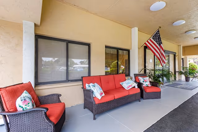 Covered front entrance porch with red-cushioned wicker seating, decorative pillows, potted plants and an American flag.