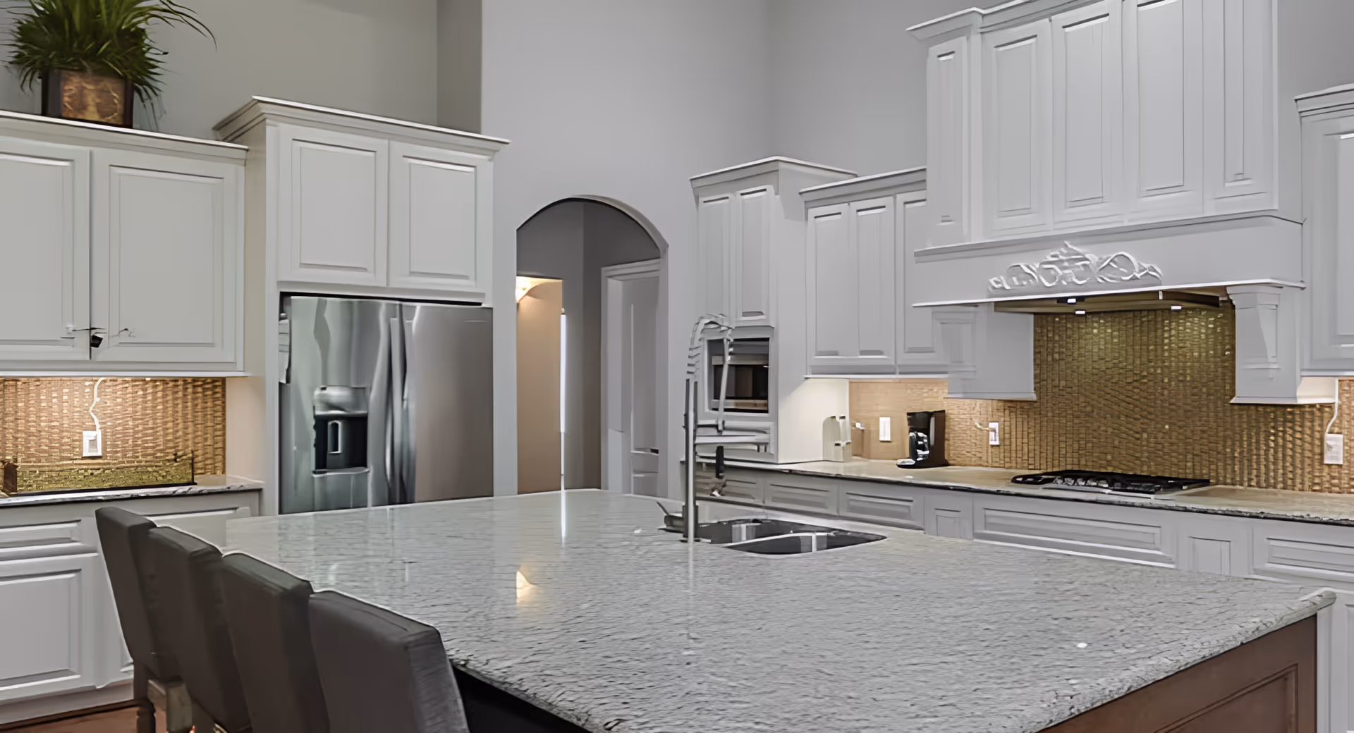 A modern kitchen with white cabinetry, a large gray granite island with a built-in sink and faucet, stainless steel refrigerator, built-in oven, and a cooktop with a decorative range hood. The backsplash features a textured golden tile design, and there are three dark-colored chairs at the island.